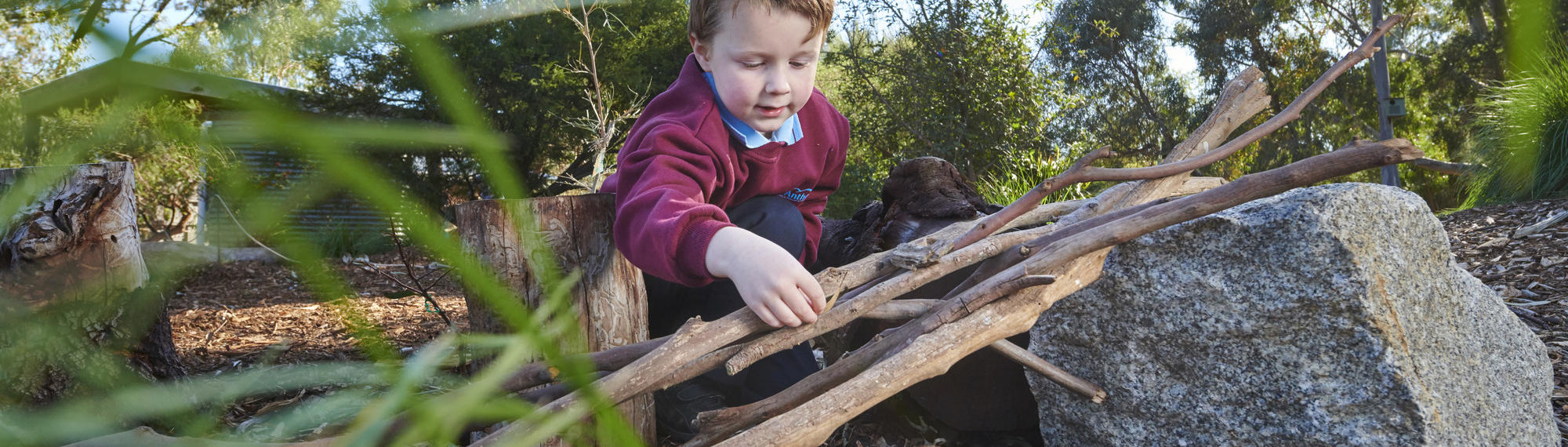 A young school student is crouched on the ground stacking some sticks