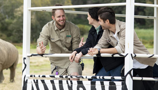 A zoo keeper stands on the back of a vehicle talking to two people. A rhino can be seen in the background.