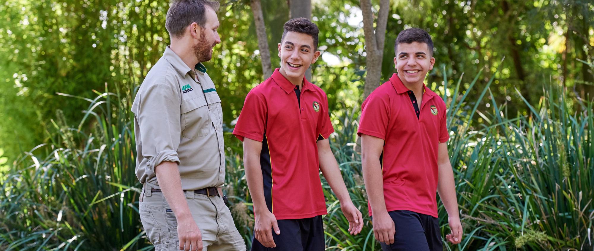 Two middle-high school aged teens in red school uniforms, walk alongside a Zoo employee dressed in khaki. 