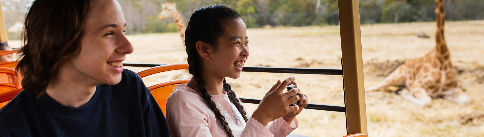 Two teens smile as they sit on a safari bus as the pass a giraffe who is sitting on the grass of the savannah