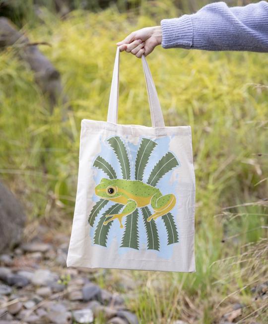 A tote bag with a bright illustration of a frog and fern leaves is held up by an arm wearing a purple knitted jumper; there is a grassy landscape in the background.