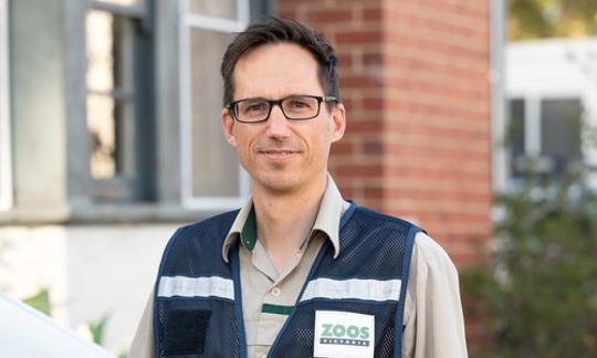 A man in glasses and a Zoos Victoria vest stares directly at the camera with a slight smile; a brick building can be seen in the background.