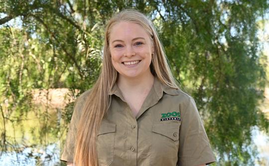 A zoo employee in uniform smiles standing in front of the wetlands 