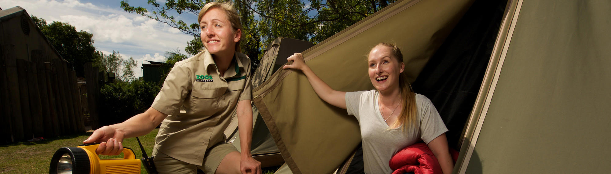 A woman lifts the door of a tent and smiles as a zoo staff members squats next to her holding a torch