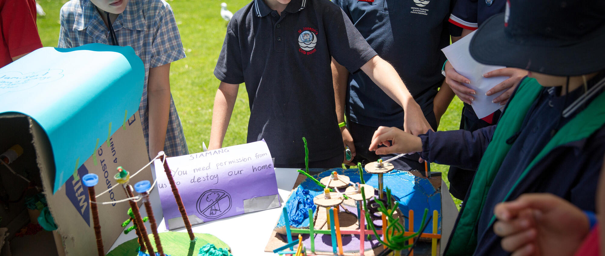 A group of primary-aged school students wearing uniform, play outside with a craft activity on a table. There is a handwritten sign on the table reading 'SIAMANG, You need permission from us to destroy our home' on it with an illustration of a tree with a cross through it.