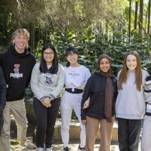 A group of nine teenage students standing in a ground and smiling for in front of a rainforest setting.