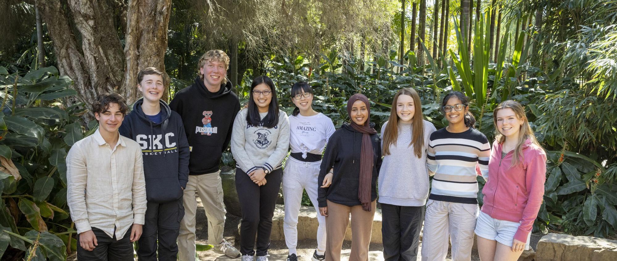 A group of nine teenage students standing in a ground and smiling for in front of a rainforest setting.