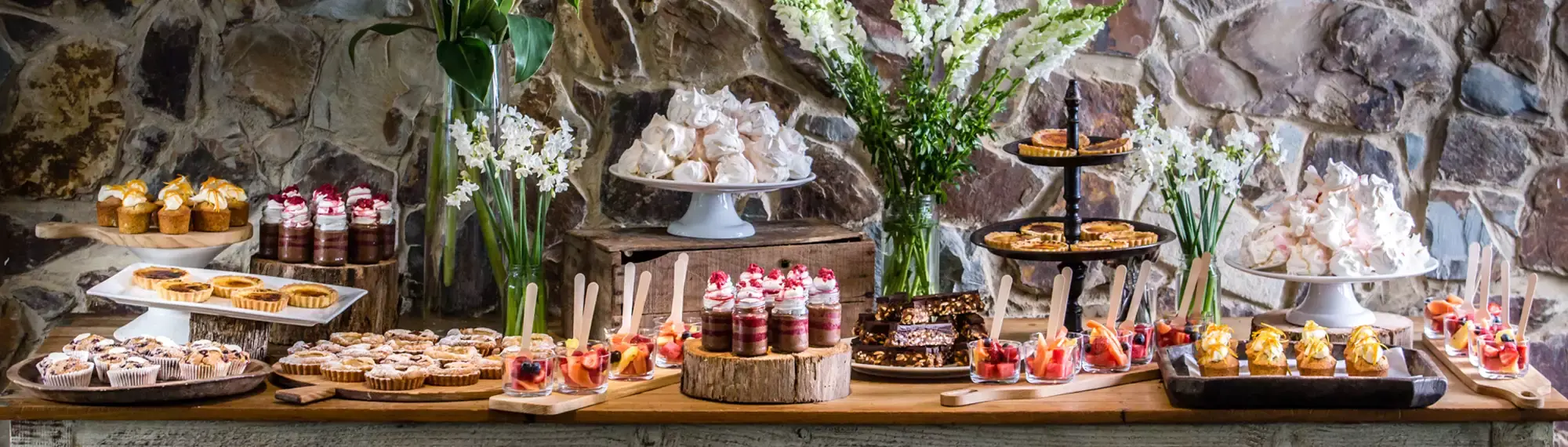 A variety of colourful sweet treats lined up on long wooden table with flowers in between the items.