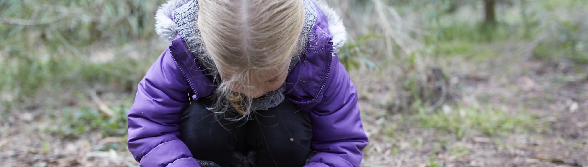 A young girl in a purple jacket bending down on the ground.